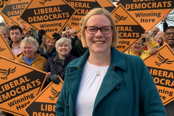 Zoe Franklin stood in front of a crowd of people. Each person in the crown is holding up a large yellow diamond-shaped sign with the words Liberal Democrats winning here